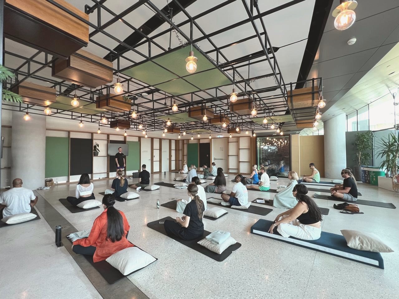 A large group seated on mats and cushions during a facilitated session in a light-filled hall