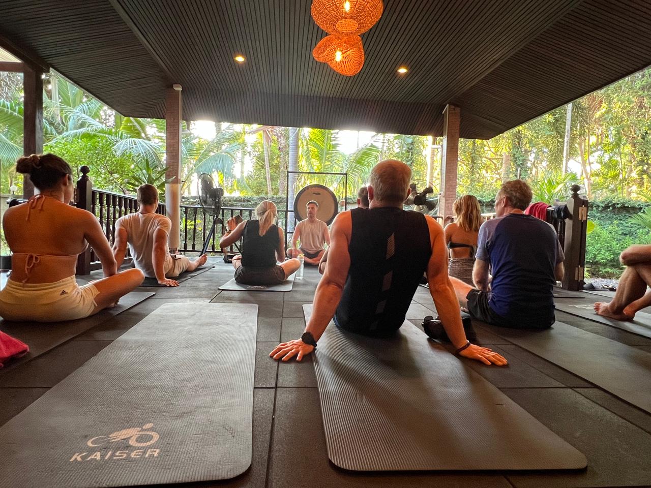 A retreat group seated on mats on an open-air veranda facing the facilitator